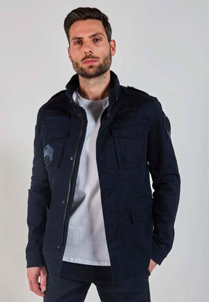 Homme aux cheveux foncés et à la barbe, portant une veste bleu marine par-dessus un t-shirt blanc, debout devant un fond gris clair uni.