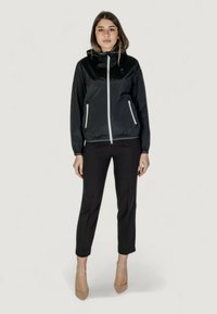 Giacca nera con zip e cappuccio, hardware argentato, polsini elasticizzati e tasche laterali. Indossata con pantaloni neri su misura e tacchi beige.