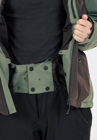 Grüne und braune isolierte Jacke mit Reißverschluss, Druckknöpfen, elastischem Bund und glatter Textur. Schwarze Hose ist darunter sichtbar.