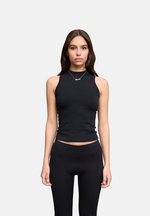 Jeune femme aux longs cheveux foncés portant un haut noir sans manches, un pantalon noir et un collier argenté, debout devant un fond clair uni.