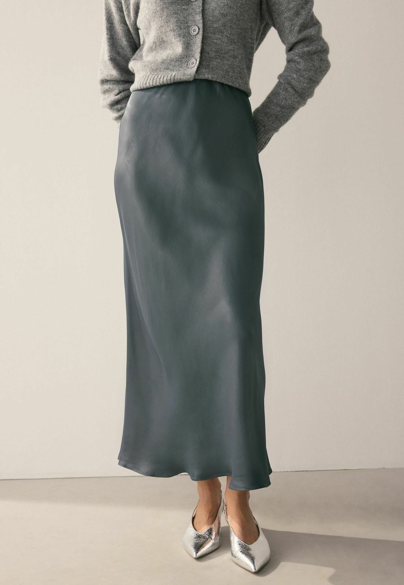 Grauer Strickcardigan über einem dunkelgrünen Satinrock mit sanften Drapierungen. Silberne spitze Ballerinas vervollständigen das Outfit.