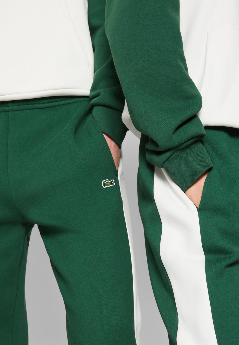 Pantaloni della tuta verdi con strisce bianche ai lati, caratterizzati da un piccolo logo Lacoste sulla tasca. Tessuto morbido, con una vestibilità rilassata e dettagli sulla tasca.