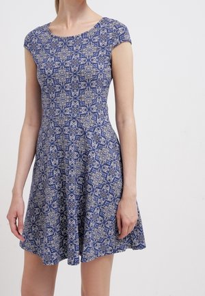 Woman wearing a short-sleeve, blue dress with white intricate floral and geometric patterns, standing against a plain light background.