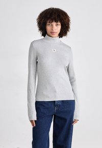Gray, long-sleeve turtleneck top in ribbed texture, featuring a small CK logo tag on the front. Paired with loose, dark blue jeans.