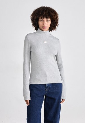 Grijze, lange mouw coltrui van geribbeld materiaal, met een klein CK-logo label aan de voorkant. Gecombineerd met losse, donkerblauwe jeans.