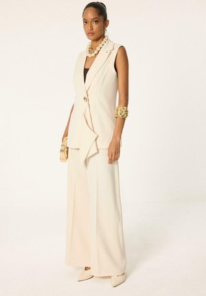 Mujer con traje sin mangas color crema con pantalones de pata ancha, collar de perlas en capas, pulsera de oro, sosteniendo un pequeño clutch dorado, de pie sobre fondo blanco.