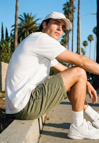 Young man in white cap, white t-shirt, green shorts, and white sneakers sitting on curb outdoors with palm trees in background.
