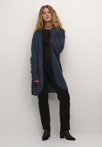 Marineblå strikket cardigan med teksturert mønster, lange ermer, åpen front, kombinert med svarte tettsittende bukser og hæler ankle boots.