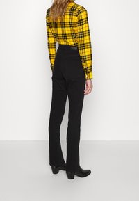 Persona che indossa una camicia a scacchi giallo e nera infilata in pantaloni neri a vita alta, con stivaletti con tacco neri, in piedi contro uno sfondo bianco.