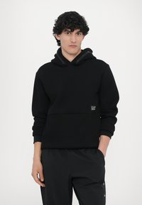 Felpa con cappuccio nera dal texture morbido, con tasca a marsupio e un patch con logo sulla parte anteriore. Abbinata a pantaloni sportivi neri.