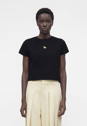 Rövid, göndör hajú nő fekete, rövid felsőt visel, amelyen kis narancssárga állatjeles embléma látható, magas derekú bézs, pliszírozott nadrággal.