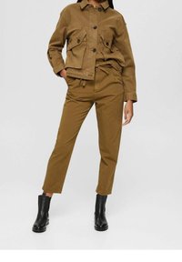 Personne portant une veste marron avec des poches à rabat, un pantalon taille haute assorti et des bottines noires, se tenant contre un fond blanc.