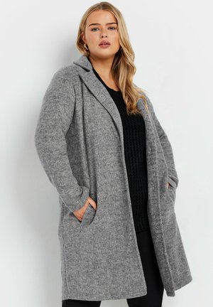 Cappotto grigio in misto lana con collo a scialle, apertura frontale e tasche laterali. Tessuto morbido e maniche lunghe, abbinato a un maglione nero a maglia sotto.