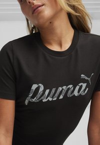  camiseta negra de manga corta, con un gran logo estilizado de "Puma" en gris con un diseño en la parte superior, cuello redondo y textura suave.