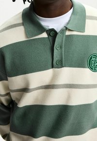 Man klädd i en grön och beige randig rugbytröja med tre gröna knappar och en rund patch-logotyp på bröstet över en vit undertröja.