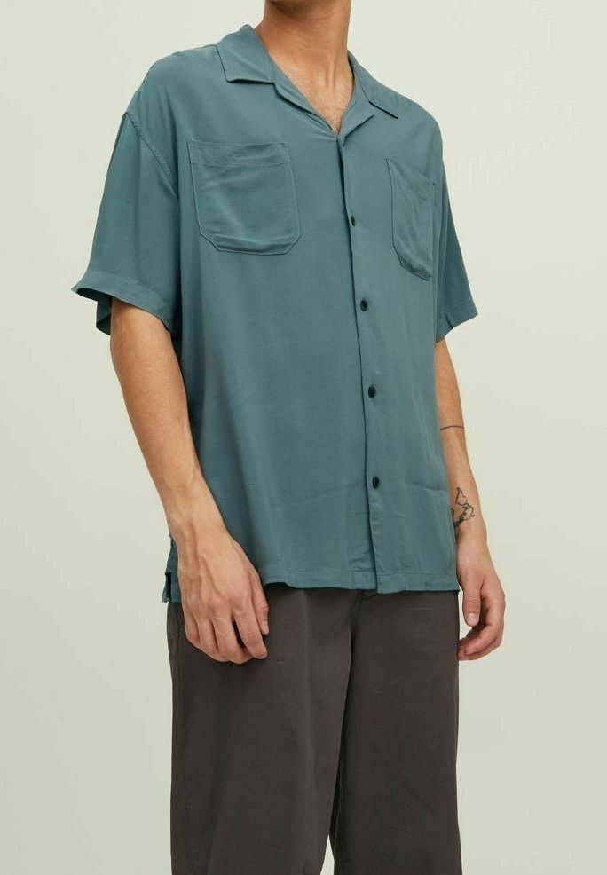 Uomo che indossa una camicia a maniche corte button-up di colore teal con due tasche sul petto e pantaloni larghi scuri, in piedi davanti a uno sfondo semplice.