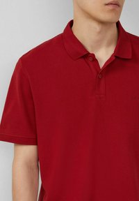 Rotes Polohemd aus strukturiertem Stoff, mit Kragen, zwei Knöpfen und kurzen Ärmeln. Dezentes Branding auf der Brust.