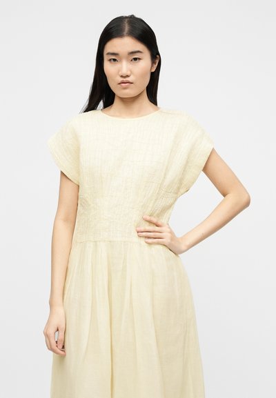 Νεαρή γυναίκα με μακριά σκούρα μαλλιά φορώντας ένα ανοιχτό μπεζ φορεματάκι με κοντά μανίκια και υφή, στέκεται με το ένα χέρι στη μέση της.