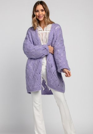 Fioletowy sweter robiony na drutach o luźnym kroju, z strukturalnym wzorem fal, otwartym przodem i opadającymi ramionami, w połączeniu z białymi spodniami.