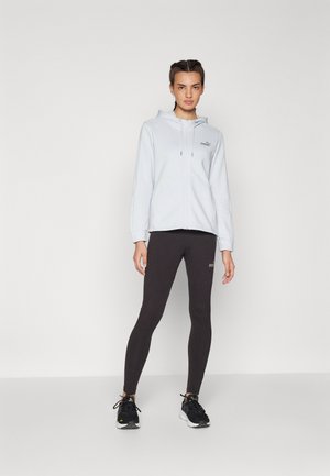 Sudadera con cremallera de color azul claro con un logo sutil, combinada con leggings negros. Presenta un corte relajado y un diseño atlético, complementado por zapatillas negras.