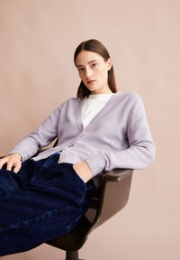Jonge vrouw met recht bruin haar zit in een stoel, gekleed in een lavendelkleurige cardigan, een wit shirt en een donkerblauwe corduroypantalon tegen een beige achtergrond.