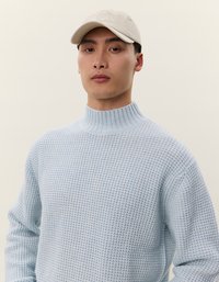 Maglione azzurro chiaro a maglia con colletto alto e motivo testurizzato, abbinato a un cappello color crema con logo. Sfondo chiaro.