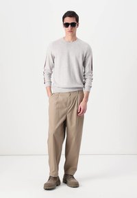 Pull gris clair avec des accents rouges sur les manches, pantalon beige ample et chaussures marron. Mannequin portant des lunettes de soleil noires, se tenant devant un fond blanc.