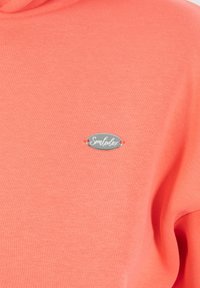 Felpa color corallo con una texture morbida e un design aderente. Presenta un logo ovale argentato con il testo "Smulodex" al centro davanti.