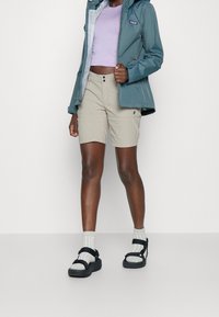 Vattentålig tealjacka, ljus lila kort topp, beiga shorts och svarta sandaler med texturerade remmar och grå strumpor.