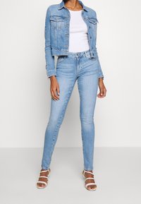 Žena v světle modré džínové bundě a džínách, s bílým přiléhavým topem a bílými páskovými podpatky, stojící před jednobarevným pozadím.