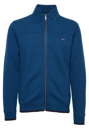 Modra zip-up jakna iz mehkega blaga, z visokim ovratnikom, navpičnimi šivi in majhnim logotipom na levi prsi. Temno modri rebrasti manšeti.