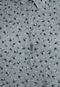 Blusa verde con bottoni caratterizzata da un motivo floreale con fiori bianchi, neri e blu. Tessuto morbido con colletto e chiusura a bottone.