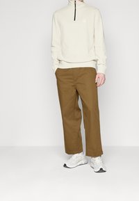 Světlý béžový svetr na zip s malým logem, kombinovaný s hnědými kalhotami s širokými nohavicemi a bílými sportovními botami. Celkově hladká textura.