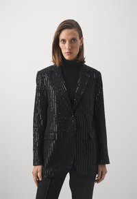 Melnā sekvīnu žakete ar vertikālu svītru rakstu, divi pogas un izteikts apkakle. Vilkta virs melna turtleneck krekla. Pielāgota piegriezuma.