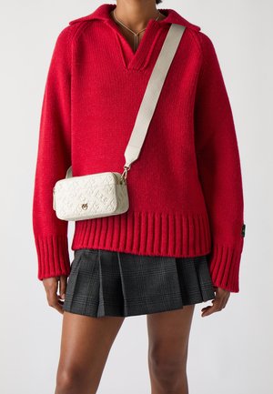 Persona que lleva un suéter de punto rojo, falda plisada de cuadros gris y un bolso bandolera blanco con un patrón en relieve.