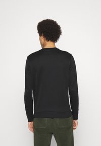 Felpa nera a maniche lunghe con collo e polsini a coste, caratterizzata da una texture liscia. Vista da dietro con pantaloni verde scuro.