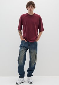 Maglietta a maniche corte color bordeaux abbinata a jeans cargo blu sbiaditi con tasche laterali. Indossata con sneakers grigie e bianche, in piedi contro uno sfondo neutro.