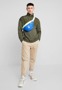 Felpa verde in tessuto con colletto zip, pantaloni beige e sneakers bianche. Viene indossata una borsa a tracolla blu e verde con dettaglio del logo.