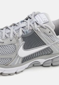 Šedá atletická bota s síťovaným svrškem, gumovou podrážkou, bílým logem Nike a texturovanými prvky pro prodyšnost a flexibilitu.