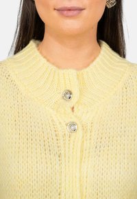 Cardigan en tricot jaune avec un col à côtes, comportant deux boutons ronds en argent et une texture douce et duveteuse.
