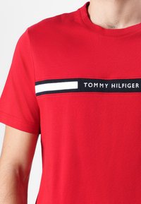 Červené bavlněné tričko s vodorovným černobílým pruhem a textem "TOMMY HILFIGER" přes hrudník. Design s krátkým rukávem.