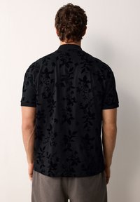 Hajlott hajú férfi fekete rövid ujjú inget visel, amely enyhe virágmintával díszített, és egy sima fehér falnak néz.