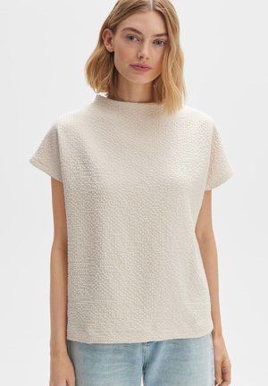 Lichte crème top met korte mouwen en hoge halslijn, met een geweven patroon en een ontspannen pasvorm, gecombineerd met lichtblauwe jeans.