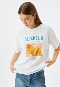 Witte katoenen t-shirt met blauwe tekst "BONJOUR" en een afbeelding van twee croissants, met een ronde hals en korte mouwen.
