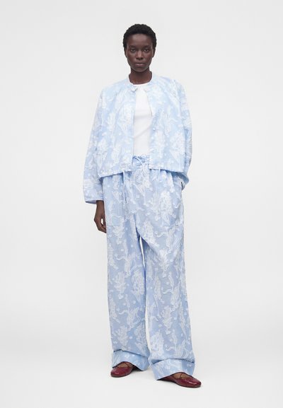 Γυναίκα στέκεται φορώντας ανοιχτό μπλε φαρδιά ζακέτα με λουλουδάτο σχέδιο και παντελόνια με φαρδιά μπατζάκια, σε συνδυασμό με λευκό τοπ και μπορντό μπαλαρίνες.