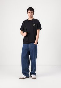 Black cotton t-shirt with a logo, paired with loose blue denim jeans and white sneakers with gray accents. Casual outfit with a shoulder bag.