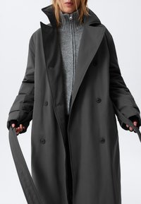 Un lungo trench coat grigio scuro con un ampio colletto, bottoni doppi e maniche larghe, indossato sopra un maglione grigio a maglia.