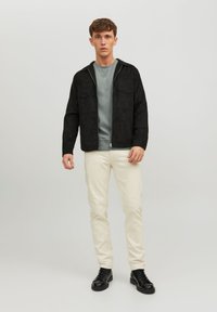 Schwarze Wildlederjacke mit zwei Brusttaschen, grauem Sweatshirt darunter und cremefarbenen Jeans. Das Model trägt schwarze Schnürstiefel. Einfarbiger Hintergrund.