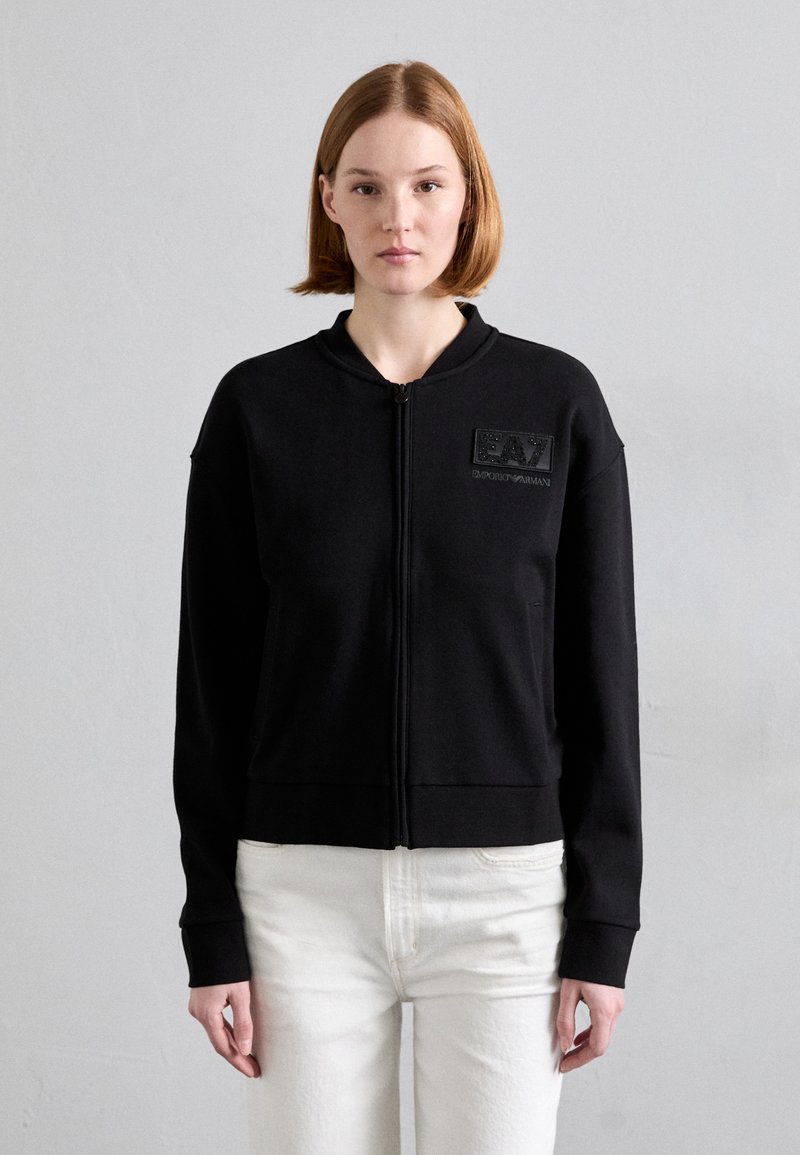 Crna zip-up dukserica s visokim ovratnikom, na lijevoj strani prsa s teksturiranim logotipom i rebrastim manžetama, u paru sa svijetlim hlačama.