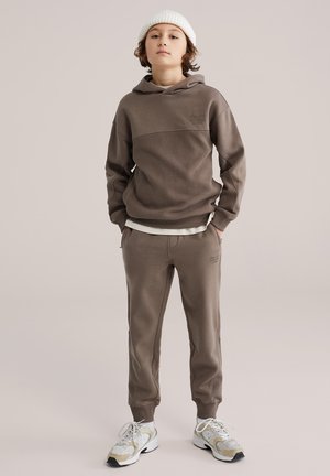 Kind met een bruine hoodie, bijpassende joggingbroek, witte muts en witte sneakers, staand met handen in de zakken tegen een effen achtergrond.